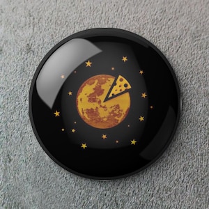 May include: A black, round button with a glossy finish. The design features a yellow moon with a slice of cheese and yellow stars on a black background. The button is a fun, whimsical accessory.