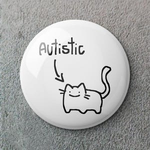 Op de afbeelding: Een witte button met het woord "Autistic" boven een zwarte cartoon kat illustratie. De kat heeft een lachend gezicht en een gekrulde staart. De button is rond en heeft een glanzende afwerking.