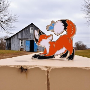 Könnte beinhalten: Ein lebendiger Emaille-Pin mit einem stilisierten Fuchs in einer spielerischen Pose. Der Fuchs hat orangefarbenes Fell, weiße Akzente und schwarze Pfoten und Ohren. Ein blauer Schmetterling ruht auf seinem flauschigen Schwanz. Der Pin ist vor einem verschwommenen Hintergrund im Freien platziert.