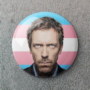 May include: A circular badge displaying a man's portrait with blue eyes, set against the transgender pride flag colours: light blue, pink, and white. The badge is on a textured grey surface. The man has a serious expression.