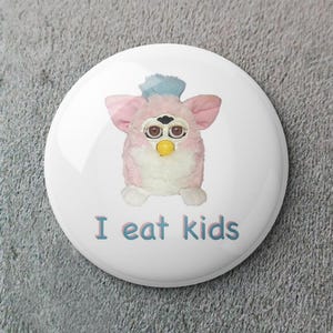 Könnte beinhalten: Ein weißer Button mit einem rosa Furby-Spielzeug mit blauen Haaren und dem Text "I eat kids" in Blau und Rosa. Das Furby hat große, runde Augen und einen gelben Schnabel.