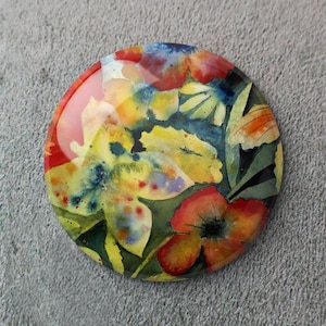 Puede incluir: Un diseño floral redondo y colorido con un acabado brillante. La obra de arte presenta flores amarillas, rojas y azules con hojas verdes, pintadas al estilo acuarela. El diseño está sobre un fondo gris.