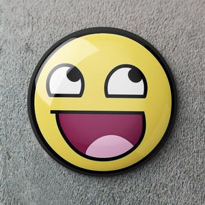 Puede incluir: Un botón redondo amarillo con un borde negro que representa una cara sonriente con los ojos hacia arriba y la boca abierta. La boca es de color rosa oscuro y los ojos son blancos con pupilas negras. El fondo es una superficie gris texturizada.