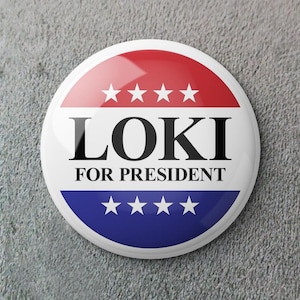 Puede incluir: Un botón redondo con un diseño rojo y azul, con el texto "LOKI FOR PRESIDENT". El botón tiene un centro blanco con la palabra "LOKI" en letras negras en negrita. Cuatro estrellas blancas están arriba y abajo del texto.