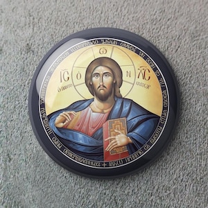 Puede incluir: Icono religioso redondo y negro que representa a Jesucristo. La imagen muestra a Jesús con un halo, sosteniendo un libro y vistiendo túnicas azules y rojas. El icono tiene texto en un patrón circular alrededor de la imagen central.