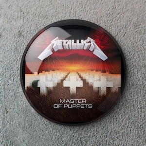 May include: A round, black button featuring the Metallica logo in white, above a cemetery scene with white crosses under a sunset. The text "MASTER OF PUPPETS" is below the image.