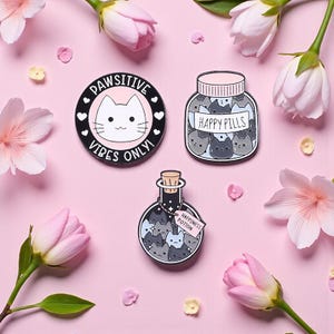 Puede incluir: Tres pines de esmalte con diseños de gatos sobre un fondo rosa con flores. Un pin dice "Pawsitive Vibes Only!" con una cara de gato. Otro dice "Happy Pills" con caras de gato dentro. El tercero dice "Happiness Potion".