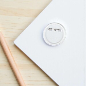 May include: A clear, round button pin with a safety pin back is centered on a white surface. The pin is partially covering the text "DC-58". A wooden pencil is in the lower left corner, and the background is a light wood grain.