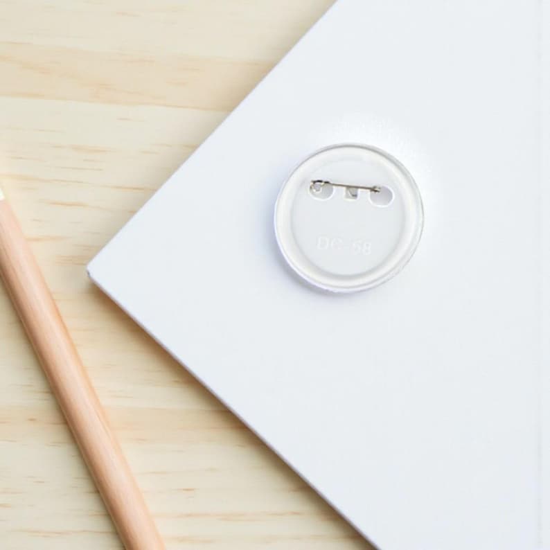 May include: A clear, round button pin with a safety pin back is centered on a white surface. The pin is partially covering the text "DC-58". A wooden pencil is in the lower left corner, and the background is a light wood grain.