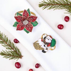 Pode incluir: Dois porta-guardanapos decorativos. Um é uma flor de poinsétia vermelha e verde com um centro de strass. O outro é um design de Papai Noel com um saco verde. Os anéis dourados estão em um guardanapo branco.