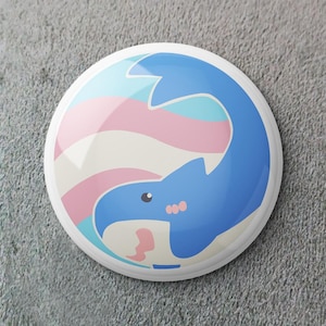 Puede incluir: Un botón redondo blanco con un diseño de ballena azul. La ballena está sobre un fondo de rayas rosas, celeste y blancas, con una pequeña forma rosa cerca de la boca de la ballena. El botón tiene un acabado brillante.