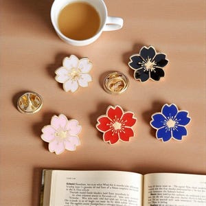 May include: A collection of flower-shaped enamel pins in various colors, including white, pink, red, black, and blue, with gold-colored centers. A cup of tea and an open book are also present.
