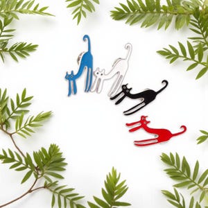 May include: A collection of cat-shaped enamel pins in blue, white, black, and red. The pins feature a stylized cat design with curved bodies and long tails. The pins are arranged on a white surface with green foliage.