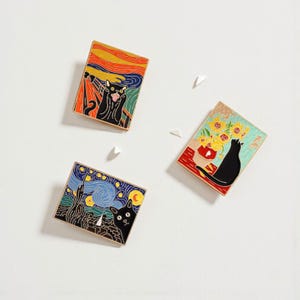 May include: Three rectangular enamel pins featuring famous art parodies. One pin depicts a black cat in "The Scream", another in "Starry Night", and the third with sunflowers. Each pin has a black border and a pin back.