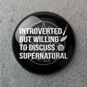 Puede incluir: Un botón redondo negro con el texto blanco "INTROVERTED BUT WILLING TO DISCUSS SUPERNATURAL". El botón presenta un pentagrama, un cuchillo, alas y un símbolo solar.
