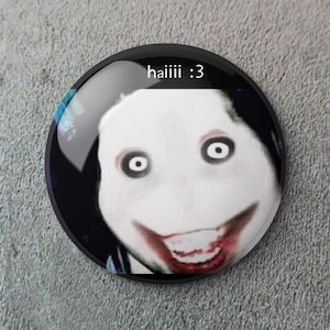 Puede incluir: Un botón redondo y negro con una cara blanca y una sonrisa sangrienta. El texto "haiiii :3" está escrito en blanco. La cara tiene ojos grandes y negros y una tez blanca.