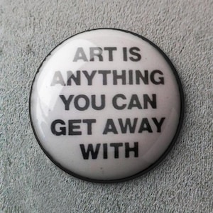 Puede incluir: Un botón redondo blanco con el texto "ART IS ANYTHING YOU CAN GET AWAY WITH" en letras negras en negrita. El botón tiene un acabado brillante y un borde negro, sobre un fondo gris texturizado.