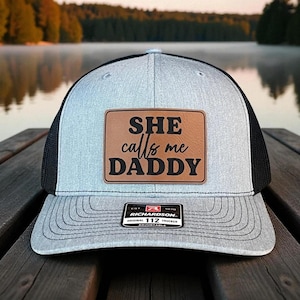May include: A gray and black trucker hat with the text "SHE calls me DADDY" on a brown leather patch. The hat has a curved brim and a mesh back. The Richardson brand label is visible.