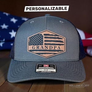 Peut inclure: Casquette de camionneur grise avec un dos en filet et un écusson en cuir avec un drapeau américain et le mot "GRANDPA". La casquette a une visière incurvée et une sangle réglable. Le mot "PERSONNALISABLE" est au-dessus de la casquette.