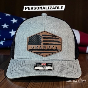 Peut inclure: Une casquette trucker grise et noire avec un écusson en cuir marron arborant un drapeau américain vieilli et le mot GRANDPA. La casquette présente un filet noir à l'arrière, un panneau avant gris et une étiquette RICHARDSON ORIGINAL 112 sur la visière.