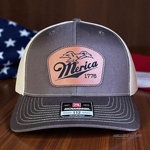 May include: A gray and tan trucker hat with a leather patch featuring an eagle and the word "Merica" above "1776". The hat has a curved brim and a mesh back. The Richardson brand label is visible.