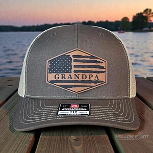Peut inclure: Casquette de camionneur grise et blanche avec un écusson en cuir marron représentant un drapeau américain et le mot "GRANDPA". La casquette a une visière incurvée et une sangle réglable. La casquette de camionneur Richardson 112 est visible.