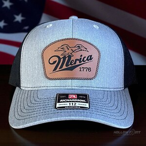 May include: Grey and black trucker cap with a leather patch displaying an eagle and the word "Merica" above "1776". The cap has a curved peak and an adjustable strap. The brand is Richardson.