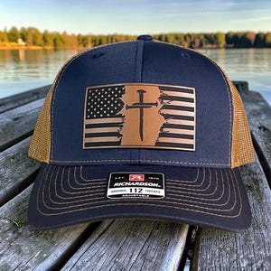 Peut inclure: Casquette de camionneur bleu marine et beige avec un écusson en cuir représentant une croix et un drapeau américain. La casquette a une visière incurvée et une sangle réglable. L'écusson indique "RICHARDSON ORIGINAL 112 TRUCKER ADJUSTABLE."