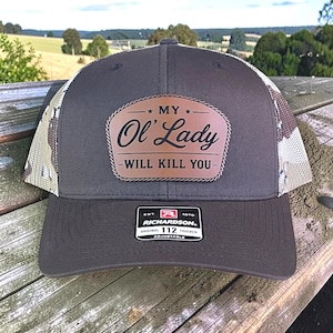 May include: A gray trucker hat with a brown leather patch that reads "MY Ol' Lady WILL KILL YOU." The hat has a mesh back and a curved brim. The brand "RICHARDSON" is visible on a tag.