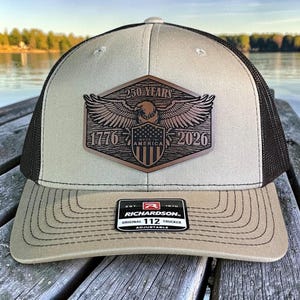 May include: Tan and black trucker hat with a patch featuring an eagle, shield, and the text "250 YEARS 1776-2026 MADE IN AMERICA." The hat has a curved brim and an adjustable strap. The Richardson label is visible.