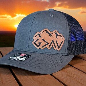 Peut inclure: Casquette de camionneur grise avec un dos en filet noir et un écusson en cuir marron représentant une chaîne de montagnes et les lettres "G > AV". La casquette a une visière incurvée et une sangle réglable.