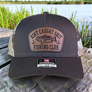 May include: A brown trucker hat with a camouflage mesh back and a leather patch that reads "Ain't Caught Shit Fishing Club" with a fish graphic. The hat has a curved brim and an adjustable strap.