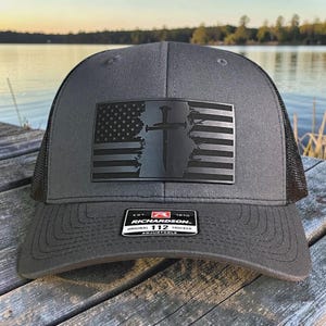 Peut inclure: Casquette de camionneur grise avec un dos en filet noir et une visière incurvée. L'avant présente un écusson rectangulaire noir avec un motif de drapeau américain et une croix. La casquette a une sangle réglable et une étiquette "Richardson 112".
