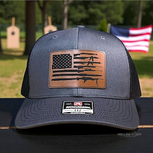 May include: Gray trucker hat with a black mesh back and a brown leather patch. The patch features a design of the American flag with guns replacing the stripes. The hat has a curved brim and an adjustable strap.