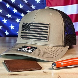 May include: A tan and brown trucker hat with a camouflage American flag patch. The hat has a mesh back and a curved brim. A brown leather wallet and an orange pocket knife are also visible.