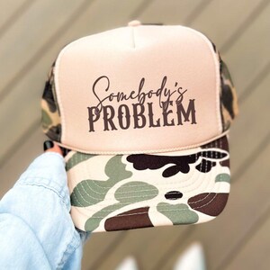 May include: A trucker hat with a tan front panel and a camouflage brim. The text "Somebody's PROBLEM" is printed in dark brown script across the front. The hat has a mesh back and a curved brim.