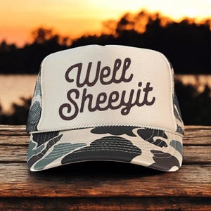 May include: A trucker hat with a white front panel featuring the text "Well Sheeyit" in a brown, cursive font. The brim and bottom panel have a camouflage pattern in shades of gray, blue, and tan. The mesh back is gray.