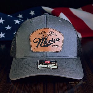 May include: A grey trucker hat with a black mesh back and an adjustable strap. A tan leather-like patch on the front features a black graphic of an eagle above the text Merica 1776. A black rectangular sticker on the brim reads Richardson 112.