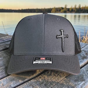 May include: A gray trucker hat with a black mesh back and a cross emblem. The hat features the Richardson 112 Trucker label and an adjustable strap. The hat is sitting on a wooden surface.