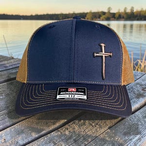 Peut inclure: Casquette de camionneur Richardson 112 bleu marine avec un dos en filet doré et un emblème de croix dorée. La casquette a une visière incurvée et une sangle réglable. L'étiquette Richardson est visible.