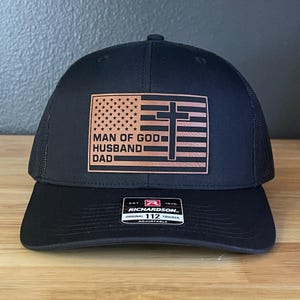 May include: Black trucker hat with a brown leather patch featuring an American flag design with a cross and the words "MAN OF GOD HUSBAND DAD". The hat is adjustable and has a mesh back.