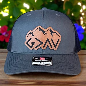 Peut inclure: Une casquette trucker gris anthracite avec un filet noir à l'arrière et un écusson en cuir beige sur le devant. L'écusson présente un motif de montagne noir au-dessus du texte G>AV. Une étiquette noir et blanc sur la visière indique Richardson 112.