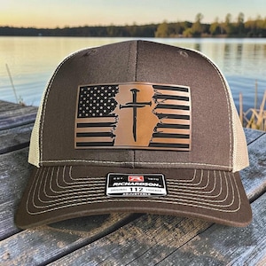 May include: Brown trucker hat with a tan mesh back and a patch featuring a distressed American flag design with a cross and nail. The hat has the text "RICHARDSON ORIGINAL 112 TRUCKER ADJUSTABLE".