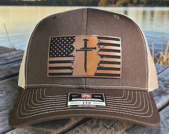Puede incluir: Gorra de camionero marrón con una parte trasera de malla beige y un parche con un diseño de bandera estadounidense desgastada con una cruz y un clavo. La gorra tiene el texto "RICHARDSON ORIGINAL 112 TRUCKER ADJUSTABLE".