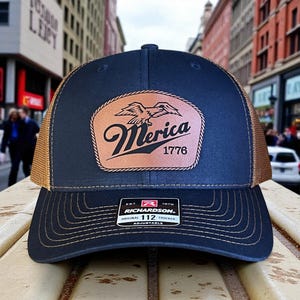 May include: Navy blue and tan trucker hat with a leather patch. The patch features an eagle design and the word "Merica" with "1776" below. The hat is adjustable and has a Richardson tag.