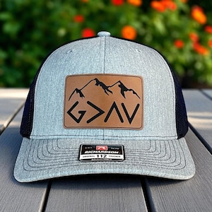 Peut inclure: Casquette de camionneur grise et noire avec un patch en cuir marron représentant un motif de montagne et les lettres "G>AV". La casquette a une visière incurvée et un dos en filet. L'étiquette de la marque Richardson est visible.