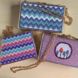 May include: Three rectangular acrylic box clutches with gold-tone chain straps. One features a purple, teal, and white chevron pattern. Another displays a pink, blue, and purple chevron design. The third has a lavender background with a floral motif.
