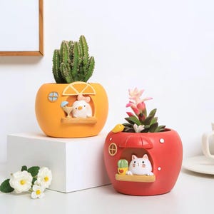 Puede incluir: Dos macetas decorativas, una naranja y otra roja, cada una con forma de fruta con una pequeña figura de animal en una ventana. La maceta naranja contiene un cactus, y la roja una suculenta.