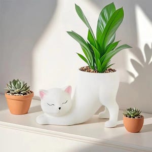 May include: A white cat-shaped planter with a green plant inside. The cat is in a stretching pose, with pink accents on the ears and nose. Two small terracotta pots with succulents are on either side.