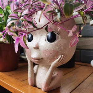 Puede incluir: Una maceta de cerámica decorativa con forma de rostro, con grandes ojos negros y detalles en rosa. La maceta está llena de una planta colgante con hojas rosas y verdes. Las manos de la persona descansan sobre sus mejillas.